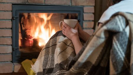 Cropped view of young adult woman covered in plaid, sitting near fireplace with fire and glass door in comfort house. Girl sitting at home with cup of beverageの写真素材