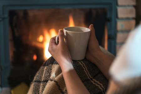 Cropped view of young adult girl with cup of tea or coffee sitting near fireplace in comfort house....