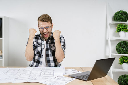 Rejoiced and happy young adult architect celebrated success, screaming, raised fists up, sitting...