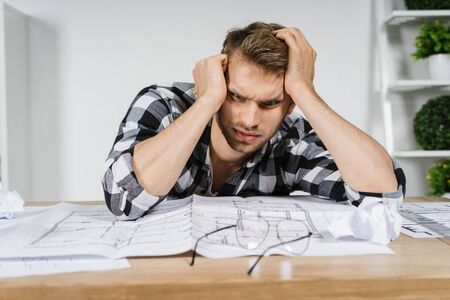 Exhausted young adult architect freelancer looking at house project, sitting behind table with tired face, working in professional bureau with blueprintsの写真素材