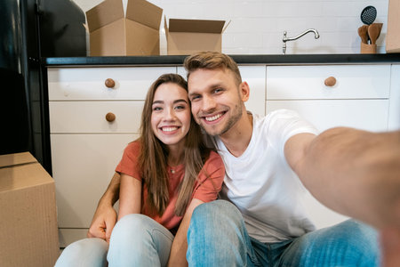 Happy young family with cardboard boxes moving in new home, sitting together on kitchen, taking self...