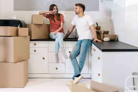 Cheerful young couple with cardboard boxes moving in new house, standing together on kitchen and...