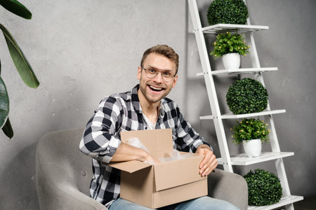 Excited and happy young adult guy spending free time at home, holding carton box in hands, unpacking...