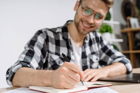Close up view of young adult freelancer using pen, writing notes in organizer, making beaming smile and sitting in workplace against copy space wall on backgroundの写真素材