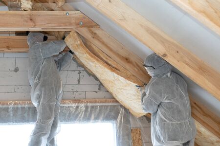 Side view of two workmen in overalls working with rockwool insulation, fasten warmth layer material on ceiling and wall, standing inside new house under constructionの写真素材
