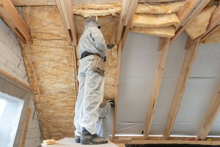 Low angle view of one foreman in overalls working with rockwool insulation material, standing inside new house under construction, fasten warmth layer material on roof ceiling and wallの写真素材