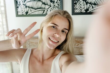 Portrait of happy young adult girl making self photo, showing victory sign,  spending morning in cozy bedroom, sitting on bed and smiling wideの写真素材
