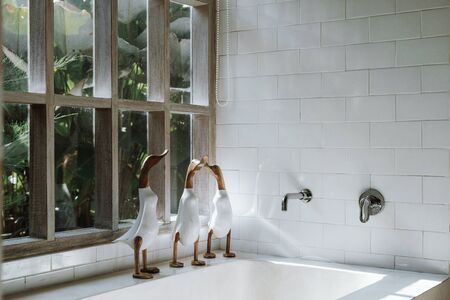 Decor with three wooden duck on top of ceramic bathtub standing near tiled wall with copy space, against window with tropical foliage plantsの写真素材