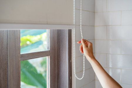Concept of lighting range control. Cropped view of woman pull rope and closed modern white jalousie...