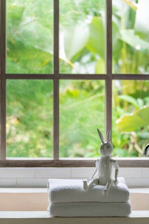 Vertical photo of clean folded towels and white decor bunny toy on top of bathtub in bathroom, against blurred window on background with tropical foliage plantsの写真素材