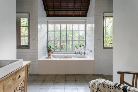 Home relaxation concept. Calm young adult woman taking bath in room with window, lying in foam...