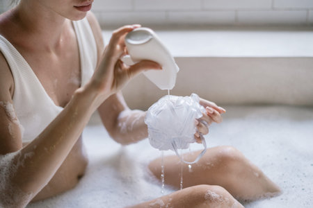 Body care concept. Cropped view of young adult woman pouring shower gel on sponge, taking bath,...