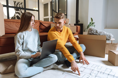 Mortgage concept. Minded young adult couple sitting in living room near architectural plan, using...