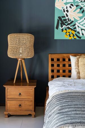 Element of cozy house with ethnic decor. Vertical photo of classic bedroom interior with lamp on wooden bedside table, comfortable bed and painting at wallの写真素材
