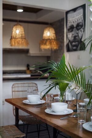 Vertical photo and selective focus on table setting in modern and cozy kitchen with white plates, utensils and tropical plants in vase at desk in dinner roomの写真素材