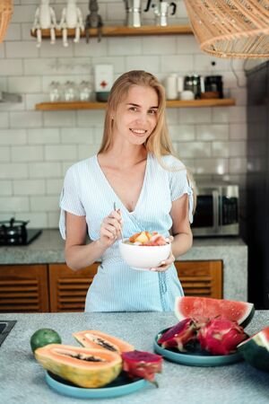 Healthy and seasonal food concept. Vertical photo of happy young adult woman standing on kitchen, eating fruit salad on breakfast, making wide smileの写真素材