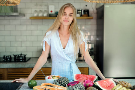 Healthy and seasonal food concept. Smiling young adult girl standing on kitchen near table with fruits, prepare cooking juicy and detox saladの写真素材