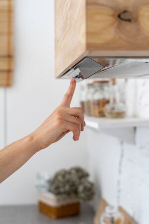 Vertical and cropped view of kitchen interior at modern house with stove hood. Woman select and switch mode at electrical panel, press on button with fingerの写真素材