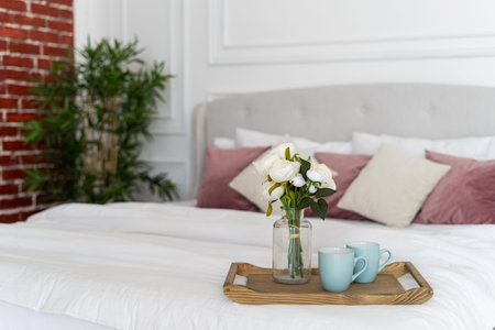 Selective focus on breakfast tray with cups and flowers in vase on comfortable soft bed at cozy bedroom. Room with light interior in modern houseの写真素材