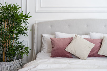 Fragment of soft cozy bed in modern bedroom with soft pillows, blanket and green plant in flowerpot. Concept of comfortable interior in roomの写真素材
