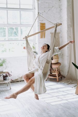 Vertical photo of carefree young adult female in bathrobe sitting on a hanging swing at home, enjoying lazy weekend morning, having fun and raised hands up, feeling freeの写真素材