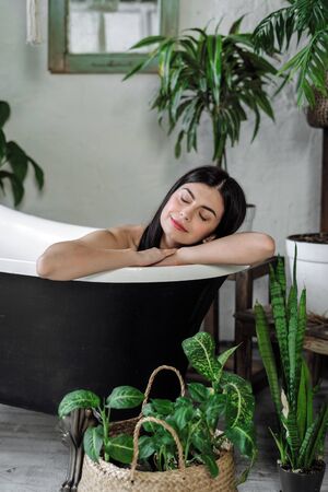 Vertical photo of smiling young adult female bathing, making dreamy face, enjoying morning routine procedure in comfortable bathroom with houseplants on backgroundの写真素材