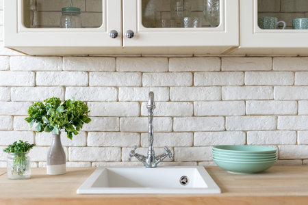 Front view of modern kitchen at home with white interior, sink and water tap, green plants and clean...