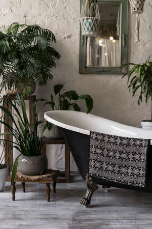 Vertical photo of elegant bathroom with stylish interior design, modern bath, textile towel, mirror on white wall and green plants in flower pots