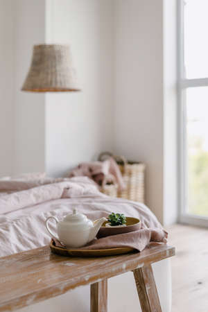 Selective focus on vertical photo of bench with home decor and teapot on tray. Morning in cozy bedroom with crumpled blanket on comfortable bed near laundry basketの写真素材