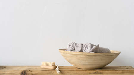 Panoramic view of clean and fresh rolled towels in wooden bowl standing on bench near soft nail brush in bathroom or spa salon. Beauty elements on table against white coy space wallの写真素材