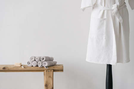 White cotton bathrobe near wooden bench with clean fresh rolled towels and nail brush. Beauty care products in bright modern bathroom or spa salonの写真素材