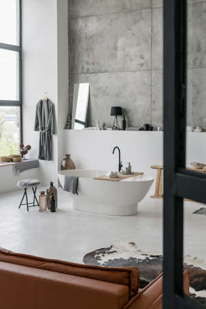 Vertical photo of modern house with bright contemporary interior design at white bathroom, comfortable ceramic bath, home decor and towels against grey concrete wallの写真素材