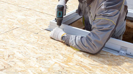 Panoramic and cropped view of professional worker using electric drill, fix the window frame for mansard room on copy space and wooden rooftop. House under construction conceptの写真素材