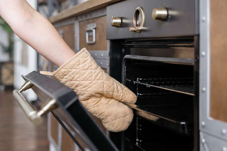 Cropped view of woman hand in kitchen glove mitt pull out tray from built in oven with open door. Cooking appliance in contemporary interior conceptの写真素材