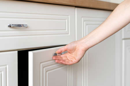Cropped view of woman hand open door at cupboard furniture standing on modern kitchen at home with white interiorの写真素材