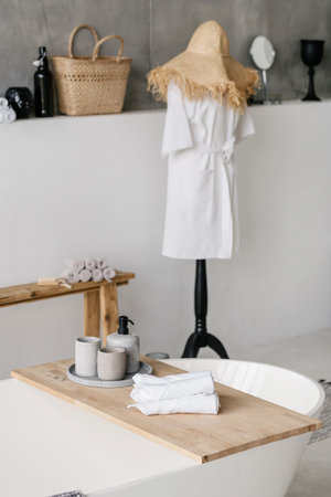 Vertical photo of white comfortable bath with clean rolled towels and house decor on wooden shelf...