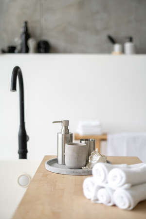 Vertical and selective focus on soap dispenser, candle, folded fresh towels at wooden shelf on white comfortable tub. Modern bathroom with house decor in contemporary interiorの写真素材
