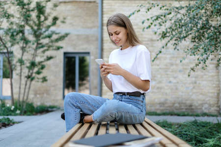 Side view of happy adult student girl using online app on modern smartphone, texting message to friends, reading news media blog, making communication on social distanceの写真素材