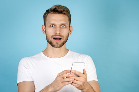 Portrait of wondered and excited adult guy in white t-shirt using modern smartphone, making steering...