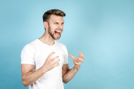 Side view of angry adult man feeling rage, screaming loud, standing isolated on blue copy space...