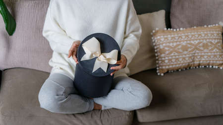 Cropped view of young adult woman holding present box in hands, sitting on comfort couch and spending weekend at homeの写真素材