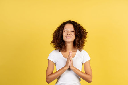 Happy and sincere adult afro american woman make prayer hands, closed eyes and standing on yellow...