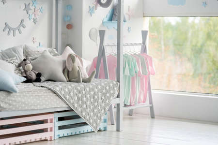 Element of childrens bedroom with plaid, pillows and toys on comfort bed, house decor, clothes on rack near window in modern apartment interior with copy spaceの写真素材
