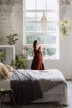 Vertical view of dreamy young woman standing near window at cozy bedroom with interior design in boho chic style. Female enjoying early weekend morningの写真素材