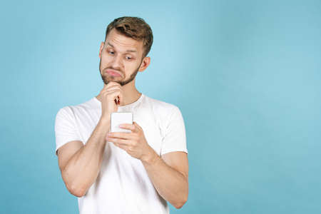 Young adult man reading interesting and not bad news on smartphone, surfing in internet, using online app, standing isolated on blue copy space background in white t-shirtの写真素材