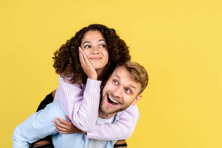 Excited young man carry on his back dreamy afro american woman, smiling wide and looking aside, standing isolated on yellow copy space backgroundの写真素材