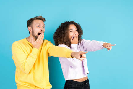 Advertising concept. Two excited and wondered mixed race friends pointing aside, smiling wide, covered mouth with palm hand, standing isolated on blue copy space backgroundの写真素材