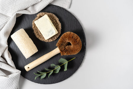 Concept of zero waste in kitchen supplies. Flat lay, top view of kitchen coconut brush, luffa sponge, solid soap, towel and eucalyptus plant on black plate stand against white copy space backgroundの写真素材
