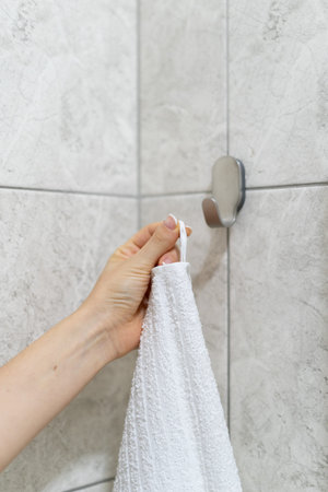 Vertical cropped view of woman hand hangs white bath towel on a hook in modern bathroom with tiled...