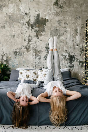 Vertical view of happy and smiling mother in pajamas lying on bed with legs up, spending lazy morning together with daughter at home. Dreamy people enjoying free time on weekendsの写真素材